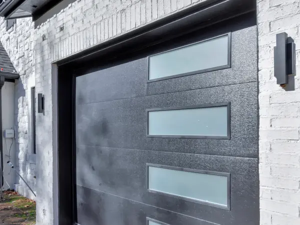 Dark glazed garage door set into painted white brick, with a slim wall light beside it